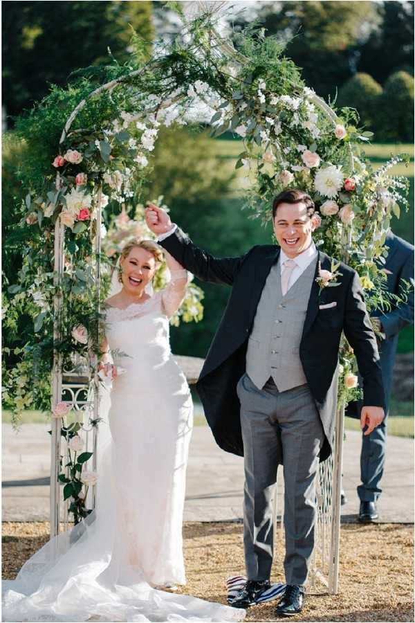 A newly married couple celebrates their just-concluded outdoor ceremony, raising their joined hands in triumph beneath a large floral arch. The arch is a white metal structure densely adorned with lush greenery, ivory and blush roses, and coral dahlias. The bride wears a long-sleeved lace ivory gown with a cathedral train and a wrist corsage, while the groom is dressed in a dark navy morning coat, a grey waistcoat, grey trousers, a blush pink tie, and a small floral boutonniere. A second male figure, likely the officiant or best man, is partially visible in the background wearing a navy suit. The setting appears to be an outdoor terrace or courtyard, with open lawns visible behind. The shot is a full-length portrait with natural afternoon sunlight.