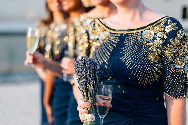 Close-up portrait of three bridesmaids standing in a row outdoors, with the foreground figure in sharp focus and the two behind progressively blurred. All three wear matching navy blue dresses with heavily embellished gold and crystal beading across the shoulders and neckline. The foreground bridesmaid holds a small dried lavender bouquet tied with twine and a champagne flute, while the others also hold champagne glasses. The styling has a formal, jewel-toned aesthetic with the gold embroidery creating a decorative contrast against the deep navy fabric.