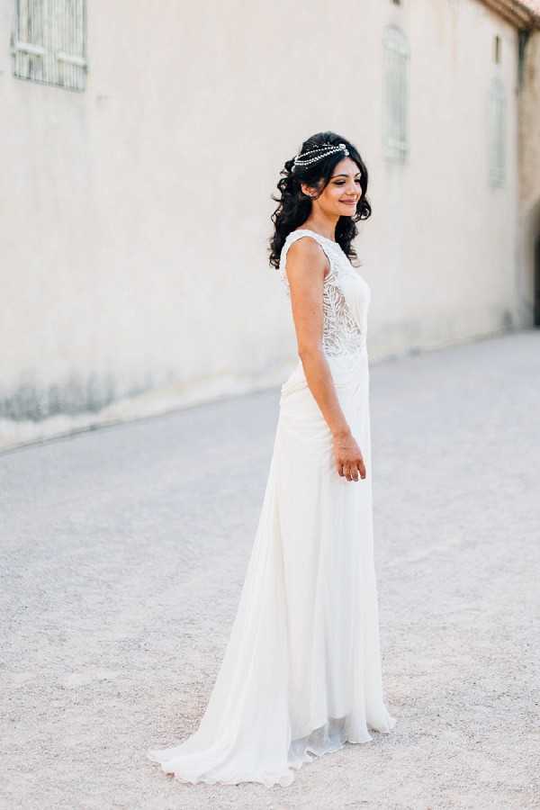 A bridal portrait of a single bride standing outdoors on a gravel surface against a pale stone wall. She wears a fitted ivory sleeveless gown with an intricate lace bodice and a flowing chiffon skirt with a small train. Her dark curly hair is worn down and accessorized with a delicate pearl or crystal headpiece draped across the crown of her head. She is turned slightly to the side, glancing back toward the camera with a smile. The shot is a full-length portrait with natural warm lighting suggesting late afternoon sun.
