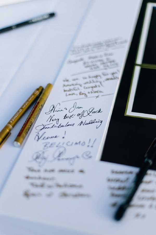 Close-up detail shot of a wedding guest book open and filled with handwritten messages from guests, including visible text such as 'Nica & Jan, Very Best of Luck, Fantabulous Wedding,' 'Hervé! BELICIMO!!' and other personal notes. Three Staedtler metallic pens in gold and black are resting on the white pages alongside the book. A black-matted frame or display board with gold trim is partially visible to the right. The overall decor palette is black, white, and gold, suggesting a classic and formal wedding styling theme.