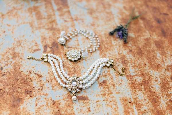A flat lay close-up of bridal jewelry arranged on a worn, rust-and-blue painted metal surface. The pieces include a multi-strand pearl bracelet with a central gold and crystal floral medallion, a pair of drop earrings featuring a crystal and pearl flower motif, and a small sprig of dried lavender placed in the upper right corner. The jewelry has a vintage-inspired aesthetic with gold-tone settings, cream pearls, and clear crystal embellishments. The composition is a detail shot with soft natural lighting.