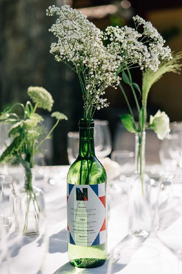 Close-up detail shot of a wedding reception table centerpiece featuring a green wine bottle repurposed as a vase, holding sprigs of white gypsophila (baby's breath). The bottle bears a custom menu label designed with a red, white, and blue geometric pattern and lists courses including Starter, Main, Selection of Cheeses, and Dessert. Surrounding the central bottle are smaller clear glass bud vases holding additional greenery and what appears to be a white bloom. The table is set with a white linen and glassware is visible in the soft-focus background. The styling is simple and rustic, with the tricolor label suggesting a French-themed or Bastille Day-inspired wedding aesthetic.
