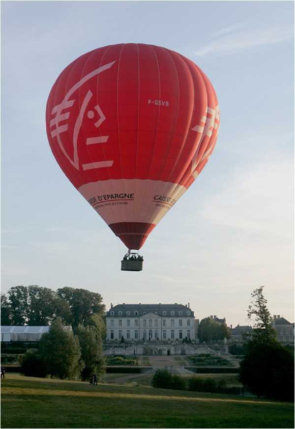 A wide-shot outdoor image showing a red hot air balloon branded with Caisse d'Epargne / Pays de Loire markings (registration F-GSVB) floating above the grounds of a classic French chateau. The balloon basket carries several passengers visible in silhouette. In the background, a symmetrical two-story French chateau with mansard roof is visible, flanked by formal hedged gardens and tree-lined grounds. A white event tent is partially visible to the left, suggesting a wedding reception setup on the property. The shot is taken from ground level in the early evening light. Potential venue feature image.