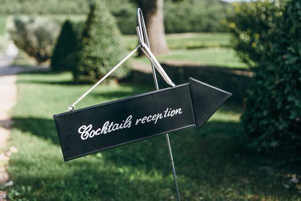 Close-up detail shot of a directional wedding sign reading 'Cocktails reception' in white cursive script on a black arrow-shaped board, suspended from a metal wire stand. The sign is positioned outdoors in a garden setting with trimmed hedges and manicured lawn visible in the soft-focus background. The signage style is classic and clean, using a black-and-white color contrast for high legibility. The composition is a tight detail shot with shallow depth of field, keeping the sign sharp against the blurred greenery.