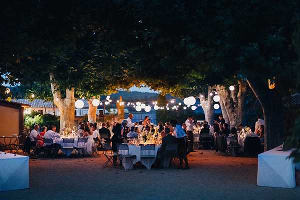 An outdoor wedding reception dinner is underway in the early evening, with approximately 50-60 guests seated at round tables arranged beneath a canopy of large mature trees. The setting appears to be a courtyard or garden of a French country property, with a low building visible to the left. White paper lanterns and string lights are strung between the trees, providing warm ambient lighting against the deep blue dusk sky. The tables are dressed with white linens and feature candlelight centerpieces, and the overall decor palette is white and natural, with a relaxed al fresco style. Wide shot taken from a low angle capturing the full reception scene.