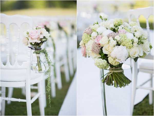 A side-by-side detail shot of outdoor ceremony aisle decor featuring white Napoleon chairs arranged in rows on a white aisle runner. The left image shows a small glass mason jar vase hanging from a chair back, containing blush pink roses and trailing greenery. The right image is a close-up of a hand-tied bouquet attached to an aisle chair, composed of white peonies, blush pink roses, cream spray roses, green viburnum snowball clusters, and dusty mauve scabiosa blooms, wrapped with twine at the stems. The overall decor palette is white, blush pink, and soft green, consistent with a classic romantic outdoor ceremony styling.
