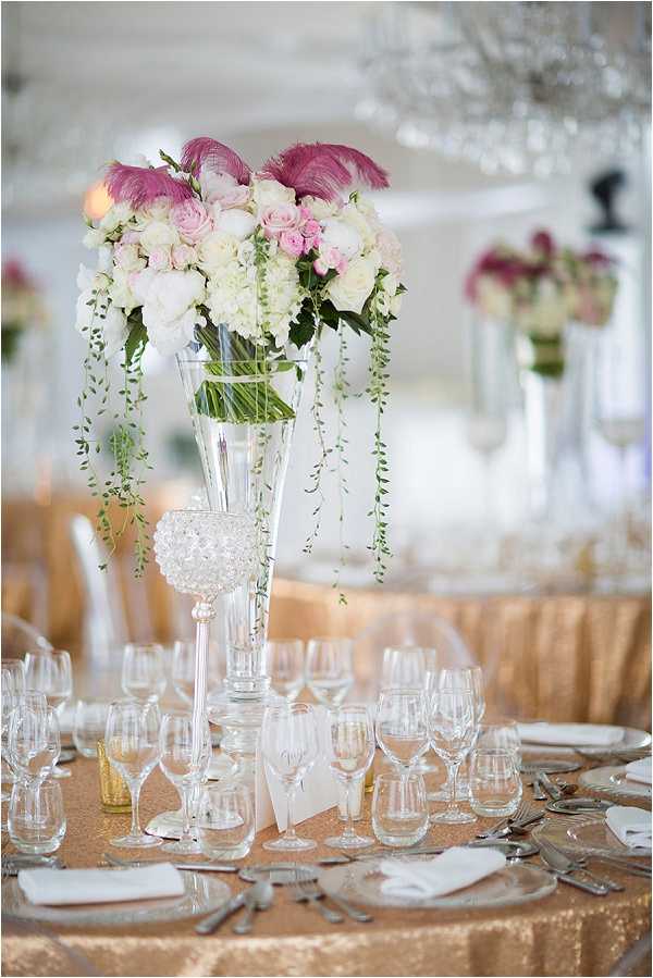 Close-up detail shot of a wedding reception table centerpiece in an indoor ballroom setting. The centerpiece features a tall trumpet-shaped clear glass vase holding a large floral arrangement of white hydrangeas, blush and light pink roses, and trailing green string-of-pearls vines, topped with deep pink feather accents. The round table is dressed in a champagne gold sequined linen with silver charger plates, multiple crystal wine and water glasses per place setting, silver cutlery, and white folded napkins. A small crystal candle holder sits at the base of the centerpiece. Additional matching tall centerpieces are visible in the soft-focus background, along with white chair covers and a chandelier, indicating a classic, formal reception aesthetic with a blush, white, and gold color palette.
