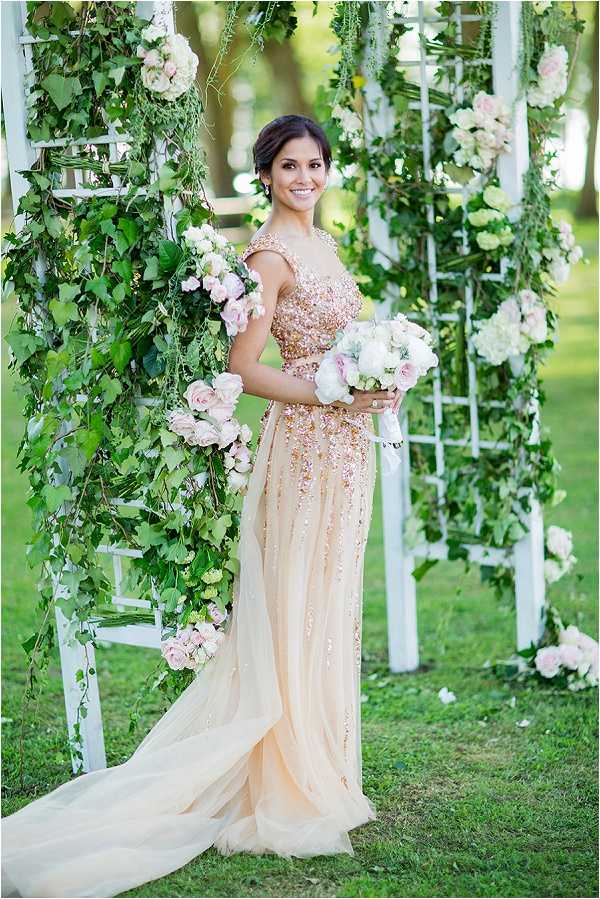 A bridal portrait taken outdoors on a lawn, with the bride standing in front of white lattice trellis structures decorated with cascading green ivy and blush pink roses. The bride wears a champagne/blush gown with a heavily embellished beaded and sequined bodice featuring rose gold and gold detailing, cap sleeves, and a flowing tulle skirt with a train. She holds a round bouquet of white peonies and soft blush roses with greenery. Her dark hair is pulled back into an updo. The styling theme is romantic garden with a soft blush and gold color palette. Medium portrait shot, full-length, with the decorative trellis framing the subject on both sides.