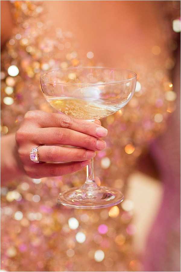 Close-up detail shot of a woman's hand holding a coupe champagne glass filled with a pale gold sparkling drink. The subject is wearing a gold and pink sequined dress that fills the background with warm bokeh reflections. A prominent pink or light purple gemstone ring, likely an engagement or cocktail ring, is visible on her ring finger. The overall color palette is warm rose gold and champagne, consistent with a reception or cocktail hour setting.