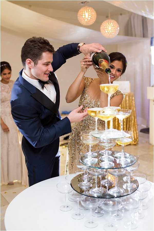 The couple is pouring champagne together into a tiered champagne tower of coupe glasses arranged on mirrored circular platforms at what appears to be an indoor reception. The groom wears a navy blue suit with a black bow tie and a gold watch, while the bride wears a full-length gold sequined gown. The reception space features warm globe pendant lights overhead and a gold sequined table covering visible in the background, suggesting a gold and navy color palette throughout the decor. A bridesmaid in a champagne or ivory lace gown is visible in the background, and the portrait-style shot captures both the couple's expressions and the champagne tower in the foreground.