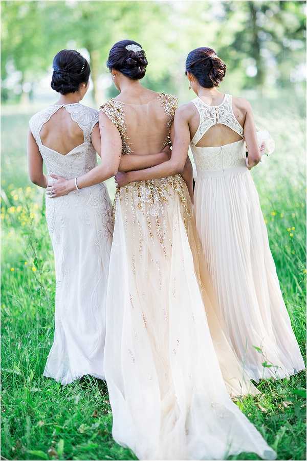 A portrait shot of three women standing outdoors in a grassy field, photographed from behind with their arms around each other. The center figure wears a floor-length champagne gown with gold and ivory beaded embellishment cascading down the back and a low open back, likely the bride. The woman on the left wears an ivory lace gown with a keyhole or cutout back detail and cap sleeves. The woman on the right wears a blush pleated chiffon gown with a lace racerback and cutout detail, and holds a small white floral arrangement. All three have dark hair styled in upswept chic updos, with the center figure wearing a crystal hair accessory. The styling is classic with glamorous embellishment details, and the shot is taken at a medium distance focusing on the backs of the gowns.