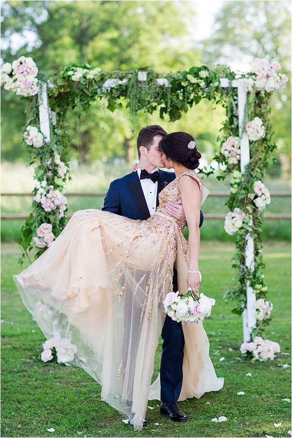 A couple shares a kiss during an outdoor wedding ceremony portrait, with the groom lifting the bride in his arms. The groom wears a navy blue tuxedo with black lapels and a black bow tie, while the bride wears a champagne and rose gold heavily embellished gown with a flowing sheer overlay skirt and a jeweled hair accessory in her upswept dark hair. She holds a small bouquet of white and blush pink roses. They are positioned beneath a white square arch decorated with lush greenery, trailing vines, and clusters of blush pink and ivory roses and hydrangeas at the corners, with white ribbon accents. The setting is an outdoor lawn area with trees visible in the background, giving the scene a garden party aesthetic. The composition is a medium portrait shot capturing both figures in full length against the arch.