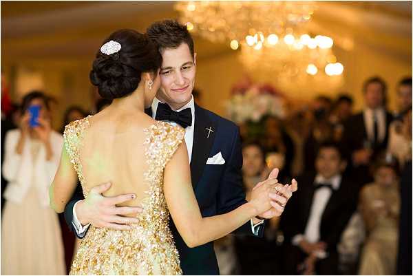 A couple shares their first dance in an indoor ballroom reception venue. The bride wears a gold heavily beaded and sequined gown with a low open back and cap sleeves, her dark hair styled in an updo with a jeweled hair accessory; the groom wears a navy tuxedo with a black bow tie, white pocket square, and a small gold cross lapel pin. A large crystal chandelier glows warmly overhead, and a crowd of formally dressed guests watches in the background, with one guest visibly photographing the moment on a smartphone. The shot is a medium portrait taken from behind the bride, capturing both faces and the detailed back of her gown, with a classic formal styling theme.