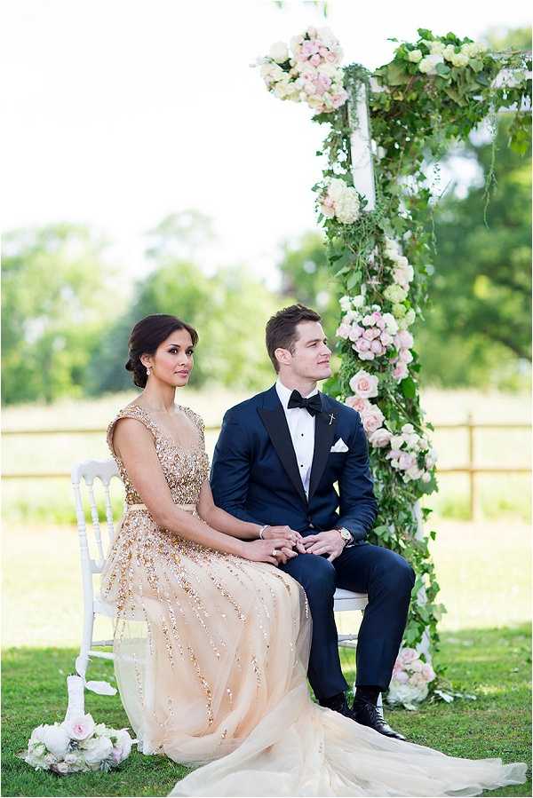 A couple is seated side by side on white chiavari chairs during an outdoor ceremony, set on a lawn with open countryside in the background. The bride wears a floor-length gold sequined gown with a flowing champagne tulle skirt, her dark hair styled in an updo, while the groom wears a navy tuxedo with black lapels and a black bow tie with a white pocket square. Behind them stands a tall white arch column heavily draped with lush greenery and clusters of blush pink roses and cream hydrangeas. A small white floral arrangement rests on the ground near the base of the chairs. The shot is a medium portrait taken at a slight distance, capturing both figures in full and the floral arch structure to the right of frame.