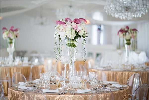 A reception room detail shot showing round guest tables dressed with gold sequin floor-length tablecloths and set with white china, crystal glassware, and silver cutlery. The centerpieces are tall clear glass trumpet vases holding arrangements of blush pink roses, white roses, pink calla lilies, and trailing greenery. Clear acrylic ghost chairs surround the tables. The indoor ballroom-style venue features crystal chandeliers and a white ceiling, with multiple identical centerpieces visible across the room in the background, creating a cohesive gold, white, and pink decor palette. Wide shot capturing the full table setting and room depth.