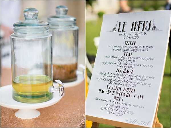 A detail shot from a French wedding reception showing two glass beverage dispensers with spigots on a white cake stand, placed on a gold sequin tablecloth, alongside a printed menu sign on a wooden easel. The menu reads 'Le Menu' for Rob and Krystal, dated 10 mai 2015, and lists courses including entrée, plat, fromage, dessert buffet, macaron wedding cake, and wines. The menu typography is styled in an Art Deco-inspired font. The setting appears to be an outdoor reception, likely a cocktail hour or dining area.