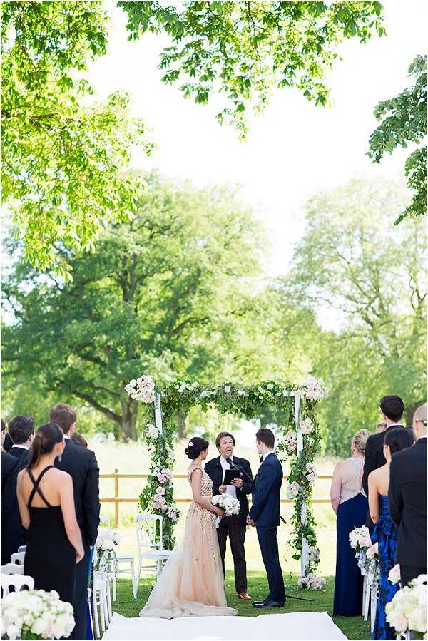 An outdoor wedding ceremony taking place on a lawn, with the couple standing before an officiant under a white square arch decorated with cascading greenery and blush pink and white florals. The bride wears a champagne-gold embellished ball gown and carries a white bouquet, while the groom is dressed in a navy suit. The bridal party flanks either side of the aisle — bridesmaids in royal blue dresses and a sequined blush dress, groomsmen in black tuxedos — with white folding chairs lining the aisle, each decorated with clusters of white and blush pink flowers. White floral arrangements also mark the aisle entrance in the foreground. The ceremony styling follows a classic palette of white, blush, and greenery with a formal, polished aesthetic. Wide shot taken from behind the seated and standing guests, looking toward the altar.