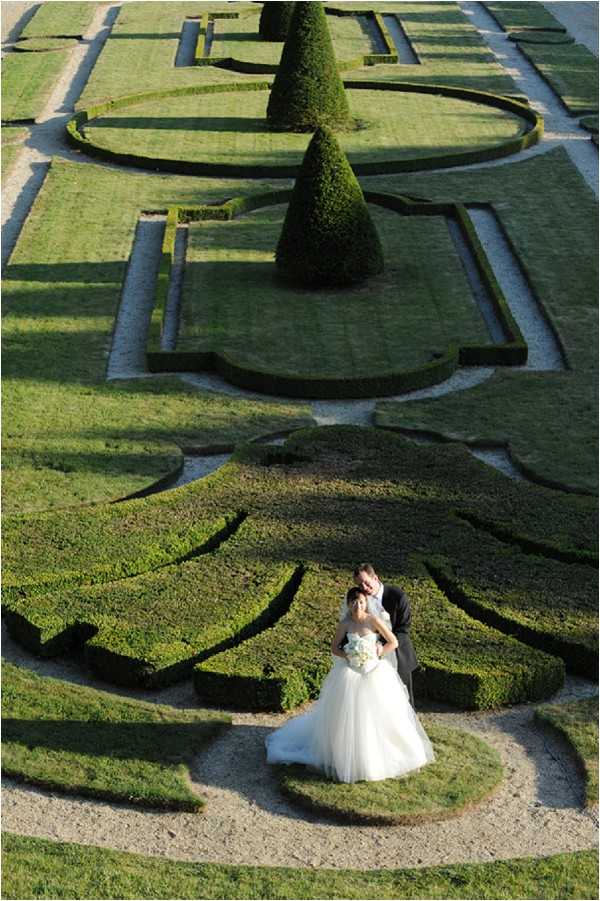 A bride and groom pose together in a formal French parterre garden, shot from a high aerial or elevated vantage point that reveals the full geometric pattern of the manicured boxwood hedges below. The groom, dressed in a dark suit, stands behind the bride and leans in close to her, while she holds a white bouquet and wears a full-skirted ivory ballgown with a strapless sweetheart neckline. The garden features precisely clipped low hedges arranged in ornate scrolling patterns with gravel pathways, and a tall conical topiary is visible in the background. The wide overhead composition places the couple as small focal figures within the grand scale of the classical garden design. Potential venue feature image.