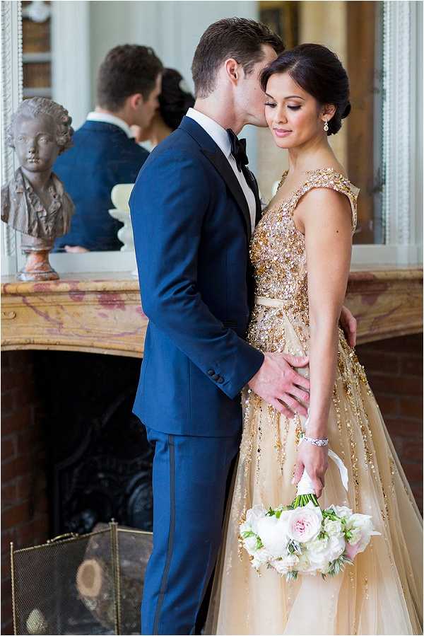 A couple portrait taken indoors beside an ornate marble fireplace mantel, with a classical stone bust sculpture visible to the left and a large mirror behind them reflecting two additional figures in the background. The groom wears a navy blue suit with black tuxedo-stripe trousers and a black bow tie, while the bride wears a gold heavily-embellished cap-sleeve ball gown with sequin and beaded detailing throughout the skirt and bodice. The bride holds a bouquet of white peonies and soft blush garden roses with white ribbon wrap, and wears a diamond bracelet and drop earrings with her hair in a low chignon. The groom leans in to kiss her temple as she looks downward. The styling is classic and formal with a gold and navy palette. Medium portrait-length shot.
