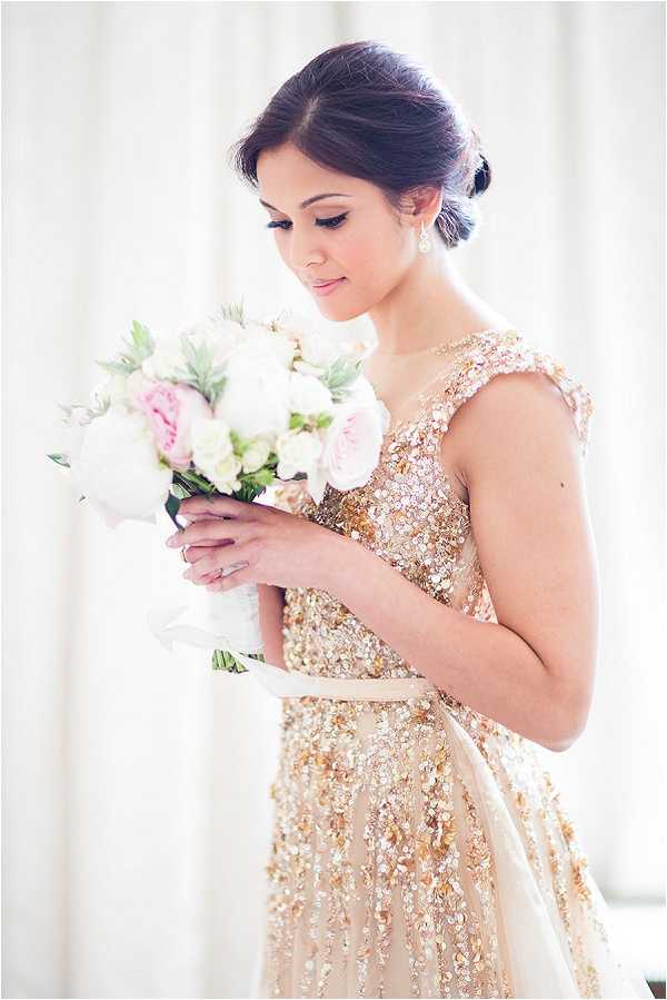A bridal portrait of a single woman wearing a gold sequined and beaded cap-sleeve wedding dress with a full skirt, photographed indoors against a soft white curtained background. She holds a round bouquet of blush pink peonies, white roses, and soft green foliage wrapped in white ribbon, and looks down at the flowers. Her dark hair is styled in a low chignon, and she wears drop earrings with natural makeup. The composition is a medium close-up portrait with a shallow depth of field and soft, diffused natural lighting, giving the image a bright, airy feel.