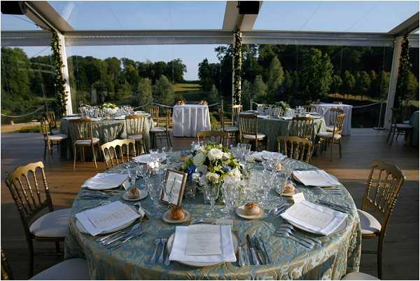 A wedding reception setup photographed from a slightly elevated angle inside a clear-roof marquee tent, with panoramic views of open parkland and trees visible through the transparent walls. The round tables are dressed in teal and gold patterned jacquard tablecloths and surrounded by gold chiavari chairs with ivory cushions. The foreground table is fully set with white plates, printed menus, multiple glasses, and silverware, centered with a low arrangement of white roses, white ranunculus, and green foliage. Additional guest tables with matching linens and floral centerpieces are visible in the background, along with a buffet or display table covered in a white cloth. The overall decor palette is teal, gold, and white with a classic, formal styling. Wide establishing shot.
