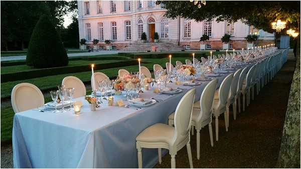 An outdoor wedding reception dinner is set up on the grounds of a French chateau, photographed at dusk in a wide shot. A single long banquet table, draped in a pale powder-blue linen, stretches the length of the frame toward the chateau's pink-rendered facade, which is lit softly in the background. The table is set with white dinnerware, crystal glassware, and tall white taper candles spaced at intervals, with low centerpiece arrangements of peach, blush, and coral flowers — likely garden roses and ranunculus — providing color accents along the runner. Cream Louis XVI-style upholstered chairs line both sides of the table, and the overall decor palette of powder blue, ivory, and peach gives the setting a classic French formal aesthetic. Potential venue feature image.