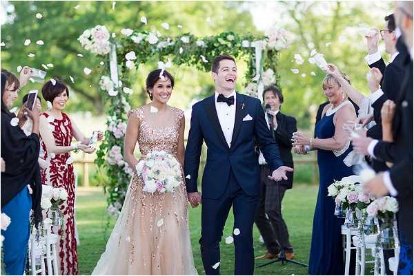 The newlywed couple walks back down the aisle during their outdoor ceremony recessional as guests on both sides toss white flower petals into the air. The bride wears a gold and rose-gold heavily embellished ball gown with a V-neckline and carries a bouquet of white and blush pink flowers, while the groom is dressed in a navy tuxedo with a black bow tie and white pocket square. The ceremony took place on a lawn with a floral arch behind them decorated with white and blush blooms and greenery, flanked by white chairs and small floral arrangements in glass vases. Guests, including women in navy and deep red dresses, smile and laugh as the couple reacts with joy; wide portrait shot.
