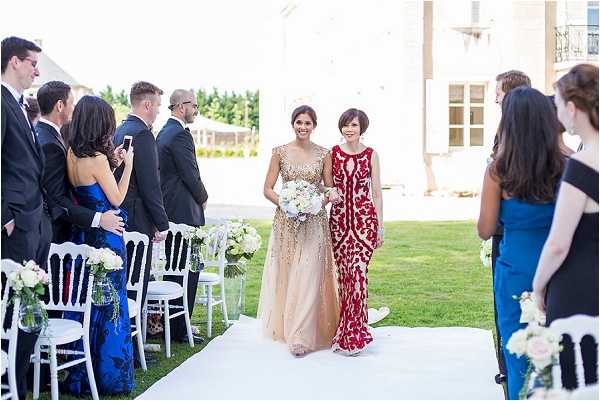 An outdoor wedding ceremony processional is underway, with a woman in a gold sequined floor-length gown carrying a bouquet of white and ivory blooms — likely peonies and roses — walking down a white aisle runner alongside a woman in a red and gold brocade floor-length dress. Guests line both sides of the aisle, seated on white Chiavari chairs decorated with small floral arrangements in mason jars featuring blush and white flowers, while additional standing guests look on; notable guest attire includes a cobalt blue gown and a royal blue dress. The ceremony takes place on a manicured lawn in front of what appears to be a French chateau or manor house, visible in the background. The overall styling is formal and classic, with a warm color palette of gold, red, and ivory, captured in a wide, eye-level shot.
