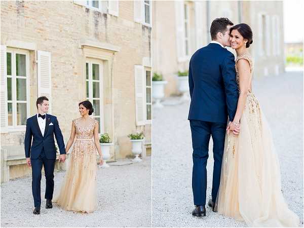 A couple portrait session split into two side-by-side images, taken outdoors on a gravel courtyard in front of a classic French chateau with pale stone facade and white shuttered windows. The bride wears a champagne-gold floor-length gown with intricate gold embroidery and a flowing tulle skirt, featuring a low open back, with her dark hair styled in an updo. The groom wears a navy blue slim-fit tuxedo with a black bow tie and white pocket square. In the left image, the couple walks hand-in-hand toward the camera smiling at each other; in the right image, the groom stands behind the bride with his arms around her as she looks back over her shoulder and smiles. Both are medium-distance portrait shots with a bright, airy, soft-light aesthetic. Potential venue feature image.