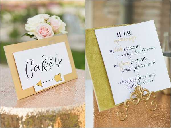 Close-up detail shot of two wedding stationery pieces displayed at a cocktail hour setup. On the left, a gold-bordered white card reads 'Cocktails' in black calligraphy with a gold arrow graphic, propped on a gold sequin tablecloth alongside a small arrangement of blush pink roses and white blooms. On the right, a bar menu card titled 'Le Bar' lists signature cocktails for the bride and groom in black and gold hand-lettered calligraphy, mounted on a gold glitter backing and displayed on an ornate gold metal easel stand. The overall decor palette is gold and white with blush accents, reflecting a classic, polished styling theme.