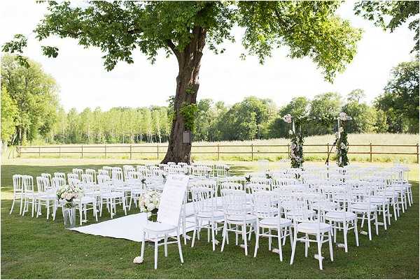 An outdoor wedding ceremony setup on a grass lawn, photographed as a wide shot before guests arrive. Rows of white Napoléon chairs are arranged in a semicircular layout with a white fabric aisle runner leading to the altar area, which is positioned in front of a large mature tree. The altar features a tall decorative arch or stand adorned with greenery and what appear to be blush and ivory floral arrangements. Small floral arrangements in blush and cream tones are placed on select chairs along the aisle. The overall decor palette is white and soft blush with green accents, giving a classic garden party aesthetic. A wooden fence and open countryside are visible in the background. Potential venue feature image.