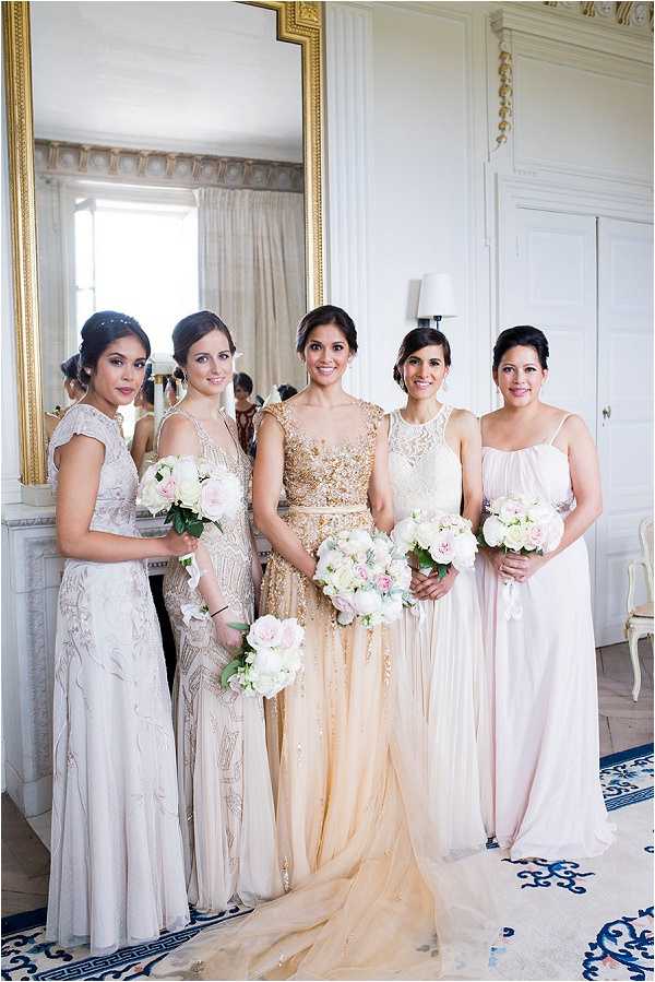 A bridal party portrait taken indoors in a classically decorated white and gold room, featuring a bride and four bridesmaids standing in front of a large gold-framed mirror. The bride wears a gold heavily embellished bodice gown with a champagne tulle skirt and holds a bouquet of white peonies and blush roses. The four bridesmaids wear mismatched floor-length gowns in a coordinated neutral palette — one silver-grey beaded gown, one champagne art-deco beaded gown, one ivory lace cap-sleeve gown, and one blush draped spaghetti-strap gown — each holding bouquets of white peonies and blush roses with greenery. The styling theme is classic with art-deco influences, and the room features ornate white paneling and a patterned blue and white rug. The image is a mid-length group portrait with soft, bright natural lighting.