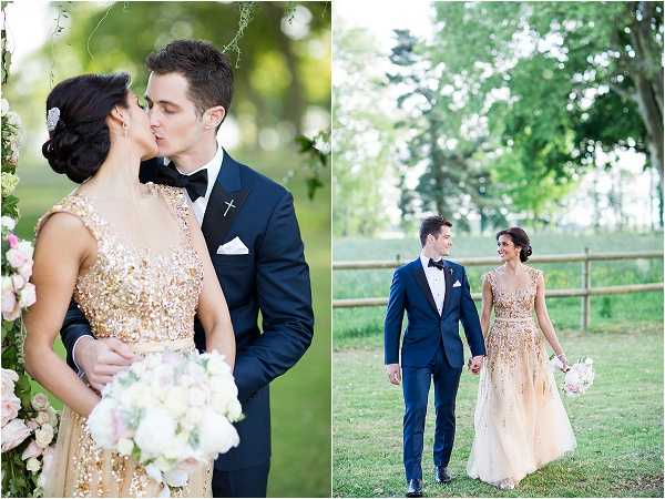 A two-panel outdoor couple portrait shoot featuring a bride and groom in a garden or countryside setting with a wooden fence visible in the background. The bride wears a gold heavily embellished cap-sleeve gown with allover sequin and beaded detailing, and carries a bouquet of white and blush blooms including what appear to be roses and greenery; her dark hair is styled in an updo with a jeweled accessory. The groom wears a navy blue tuxedo with black bow tie, black lapels, and a white pocket square, with a small cross lapel pin. In the left panel, the couple shares a kiss beside a floral arrangement of blush and white flowers with trailing greenery; in the right panel, they walk hand in hand smiling at each other in a wider portrait shot. Both panels are brightly lit with soft natural light, giving a clean, classic editorial feel.