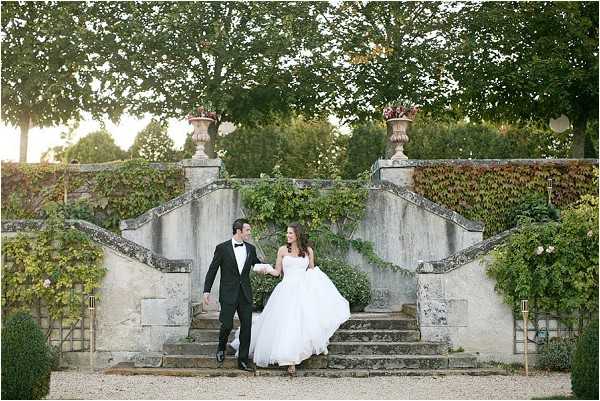 A couple portrait taken outdoors on a gravel path at the base of a formal stone double staircase covered in ivy, with large ornamental stone urns at the top balustrade. The groom wears a black tuxedo with white dress shirt and black bow tie, while the bride wears a strapless white ballgown with a full tulle skirt; they are holding hands and looking at each other as they walk. The setting has a classic French formal garden style, with warm golden late-afternoon light visible through the trees behind them. Wide shot, full-length portrait composition. Potential venue feature image.