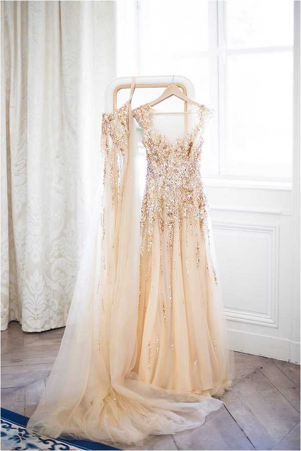 A detail shot of a bridal gown hanging on a white and gold garment rack in front of a bright window in an indoor getting-ready space. The dress is a champagne/blush tulle ball gown heavily embellished with gold and silver sequin and beaded detailing across the bodice and cascading down the skirt, with a matching heavily embellished cape or sleeves draped over the hanger. The floor beneath is herringbone parquet wood and a patterned white damask curtain is visible to the left. The image is well-lit with natural backlight, creating a bright, airy atmosphere around the dress.