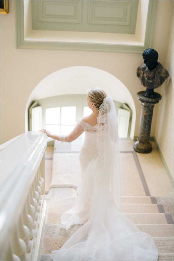 A bridal portrait taken indoors on a grand staircase of what appears to be a chateau or manor house. The bride stands with her back to the camera, resting one hand on a white painted balustrade, wearing a fitted ivory lace gown with long lace sleeves and an off-the-shoulder neckline, with a cathedral-length veil trailing down the stairs. Her blonde hair is styled in an updo. The interior features cream-painted walls with molded ceiling details, a stone-tiled landing floor with a geometric border pattern, an arched alcove with a window providing soft backlight, and a dark bronze classical bust on a marble pedestal column. The composition is a three-quarter-length portrait shot from slightly above and behind the bride, giving a clear view of the gown's train and veil length. The overall decor style is classic French formal interior. Potential venue feature image.