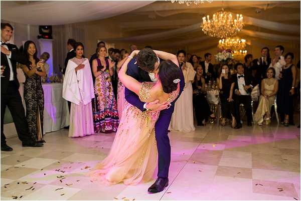 The couple shares their first dance inside a tented or marquee reception venue, with the groom dipping and kissing the bride at the climax of the dance. The bride wears a gold and blush heavily embellished gown with a flowing skirt, while the groom is dressed in a navy blue suit. Gold and orange confetti is scattered across the white checkered dance floor, and pink uplighting illuminates the floor area. Guests, numbering approximately 40-50 visible, line the perimeter of the dance floor watching and applauding, dressed in formal black-tie attire including several women in floor-length gowns; one bridesmaid or guest wears a colorful printed full-length dress. The venue interior features crystal chandeliers and draped white ceiling fabric. A priest or officiant in white robes stands among the guests to the left. The shot is a wide-angle candid taken from a slightly elevated angle capturing the full dance floor scene.