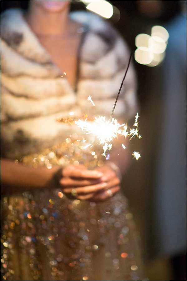 A close-up detail shot of a woman holding a lit sparkler at what appears to be an evening wedding reception. The subject is wearing a gold sequined skirt and a white or cream faux fur stole, suggesting a glitzy, glamorous party atmosphere. The sparkler is the focal point of the image, with the background and subject intentionally blurred through shallow depth of field, creating warm bokeh light spots in the background. The overall color palette is warm gold and ivory.