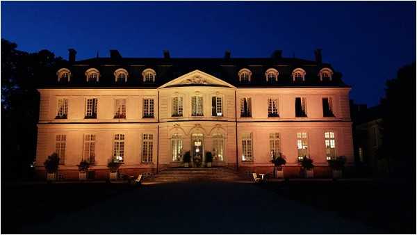 Exterior night shot of a large French chateau, photographed from ground level in a wide-angle composition. The classical 18th-century style building features a symmetrical facade with dormer windows on the upper floor, tall French windows on the lower floors, and a central entrance with a stone staircase and ornate pediment. The building is lit with warm amber uplighting against a deep blue dusk sky, and interior lights glow through the windows, suggesting an active event inside. Potted topiaries flank the entrance steps and ground-floor windows. No people are visible in the frame. Potential venue feature image.