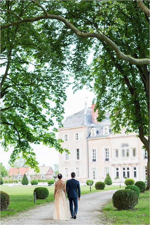 A couple walks together along a gravel path leading toward a French chateau, photographed from behind in a wide portrait shot. The bride wears a champagne-colored gown with gold embroidered detailing across the open back, and her hair is styled in an updo with a small hair accessory; the groom wears a navy suit. The chateau is a pale stone, multi-story classical French manor with a slate mansard roof, ornate window surrounds, and a cupola, set within formal grounds featuring clipped topiary spheres and cones. Potential venue feature image.