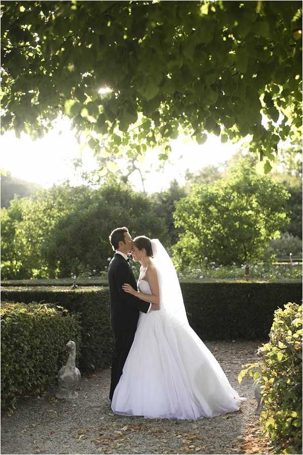 A couple shares a kiss during an outdoor portrait session in a formal garden setting, standing on a gravel pathway flanked by neatly trimmed low hedgerows and a stone duck sculpture. The bride wears a white strapless ballgown with a full tulle skirt and a long cathedral-length veil, while the groom is dressed in a black tuxedo with a white shirt. The shot is backlit by warm late-afternoon sun filtering through a canopy of tree branches overhead, creating a bright, glowing background. The composition is a full-length portrait taken at medium distance, capturing both figures and the surrounding garden landscape.