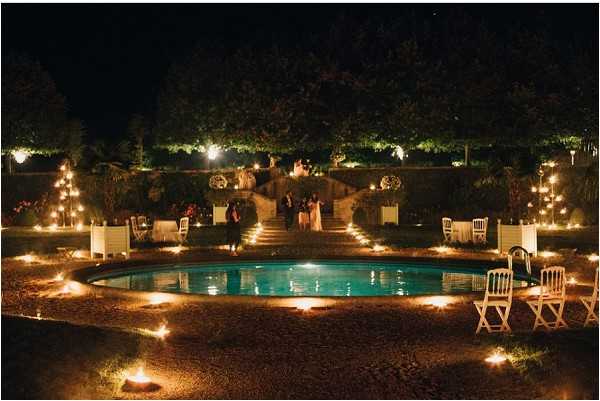 An outdoor evening wedding reception photographed from an elevated wide-angle perspective, centered on an oval pool surrounded by candles and small ground-level lanterns placed along the pool's perimeter. The venue features a formal garden layout with stone steps leading up to a lit terrace area where a small group of approximately 8-10 guests are gathered. White folding chairs are arranged around the pool area, and the scene is lit almost entirely by warm candlelight, ground torches, and decorative tiered candelabras or light trees flanking the steps on both sides. The overall decor palette relies on warm amber lighting against the dark night sky, creating a candlelit classical garden atmosphere. Potential venue feature image.