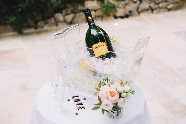 A close-up detail shot of a cocktail hour champagne display set on a white linen-covered table outdoors on a stone terrace. A large acrylic ice bucket filled with ice holds a bottle of Jecder champagne, flanked by two tall acrylic champagne flutes. In the foreground, a small glass bud vase holds a mini arrangement of blush pink roses and white buds with greenery, alongside wooden letter tiles spelling 'LOVE'. The overall decor palette is white, clear acrylic, and blush pink, giving a modern and clean aesthetic.