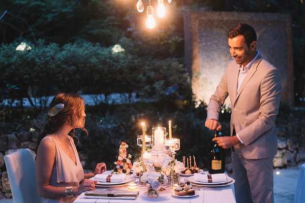 A couple shares an intimate outdoor dinner setting at dusk, with the groom in a light gray suit opening a bottle of wine and the bride seated at a white-linen table wearing a pale gray sleeveless dress with a floral hair accessory. The styled table features a centerpiece of pillar candles in glass holders, small floral arrangements with blush and coral blooms, and neatly folded white napkins on white plates with dark ribbon ties. Overhead Edison bulb pendant lights provide warm ambient lighting alongside the candlelight, creating a moody evening atmosphere. The scene appears to be a styled editorial or intimate reception setup in an outdoor garden or terrace setting, captured as a medium-wide portrait shot.