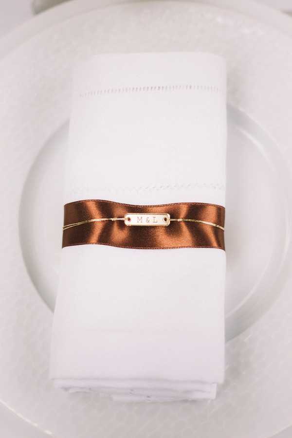 Close-up detail shot of a folded white linen napkin with hemstitch embroidery, placed on a white ceramic plate. The napkin is wrapped with a deep copper-brown satin ribbon tied around a delicate gold chain bracelet bearing a small gold rectangular charm engraved with the initials 'M & L', serving as a personalized wedding favor doubling as a napkin ring. The overall table setting palette is white and warm gold with deep copper-brown accents, reflecting a classic and refined reception styling approach.