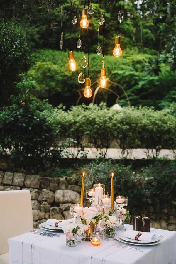 An outdoor reception tablescape set for two in a garden setting at dusk. The rectangular table is dressed in a white linen tablecloth and set with white plates, silver flatware, and white cloth napkins with copper-toned napkin rings. The centerpiece features a cluster of gold taper candles, glass pillar candle holders, floating candles, and small votive candles alongside low arrangements of white and blush flowers with greenery. Overhead, a hanging installation of Edison-style exposed filament bulbs mixed with crystal pendants provides warm ambient lighting. The overall decor palette is white, gold, and copper with a modern-romantic styling. Wide shot, slightly elevated angle.