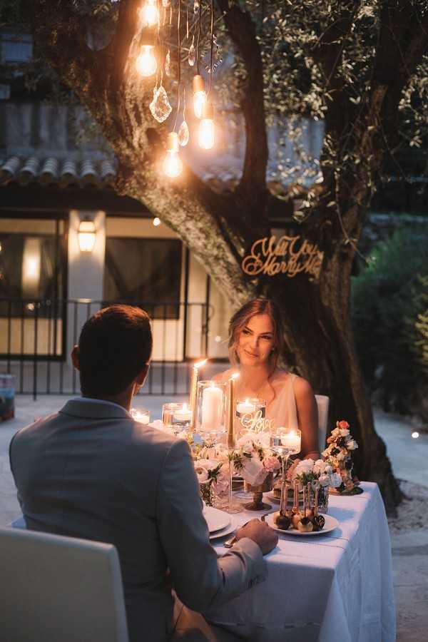 A couple sits together at an intimate outdoor sweetheart table set for two in a courtyard at dusk, with a Provençal-style stone building visible in the background. The table is dressed in a white linen cloth and styled with tall glass pillar candle holders, low arrangements of blush and white florals, and small taper candles in gold holders, creating a warm candlelit atmosphere. Hanging above the table from a tree are Edison bulb pendant lights with crystal or geode accents, and a gold cursive 'Will You Marry Me' sign is mounted on the tree trunk behind them. The man, seen from behind in a light grey suit, faces a woman in a white sleeveless dress whose face is softly lit by the candles; the composition is a medium portrait shot taken from slightly behind and above the groom's shoulder.