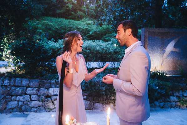 A couple shares a joyful, animated moment outdoors in a garden setting at dusk or evening, both laughing and gesturing expressively toward each other. The woman wears a blush-pink short dress with a dark draped scarf or wrap, with her hair loosely styled up, while the man wears a light beige suit with an open-collar white shirt. Several lit candles are visible in the foreground at low level, providing warm candlelight against the cool blue ambient lighting of the evening. The shot is a medium portrait framed at mid-distance, with a stone wall and sculpted garden feature visible in the background.