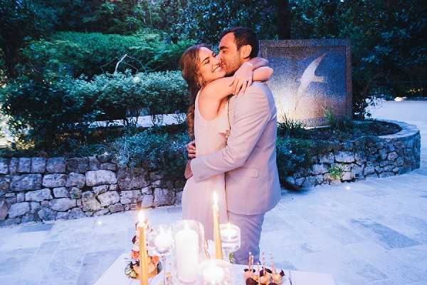 A couple shares a kiss outdoors in the early evening at what appears to be a private terrace or garden setting, with a decorative stone wall and abstract sculpture visible in the background. The groom wears a light beige suit and the bride wears a sleeveless blush or ivory slip-style dress. In the foreground, a small table holds lit taper candles, a white pillar candle, and what appear to be small desserts or appetizers, suggesting an intimate dinner or cocktail moment. The shot is a medium portrait framing the couple from the waist up, with warm candlelight contrasting against the cool blue dusk light.