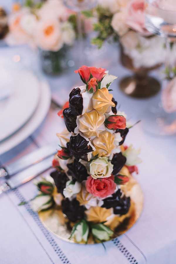 Close-up detail shot of a croquembouche-style tower dessert displayed on a gold cake board at a wedding reception table. The tower is constructed from dark chocolate-dipped profiteroles and gold and cream-colored meringue kisses, decorated with coral, ivory, and blush mini roses and small flower buds tucked throughout. The dessert sits on a white linen tablecloth with a subtle woven border pattern, and the background shows blurred place settings with silver flatware, white plates, and floral centerpieces featuring peach and blush roses with greenery.